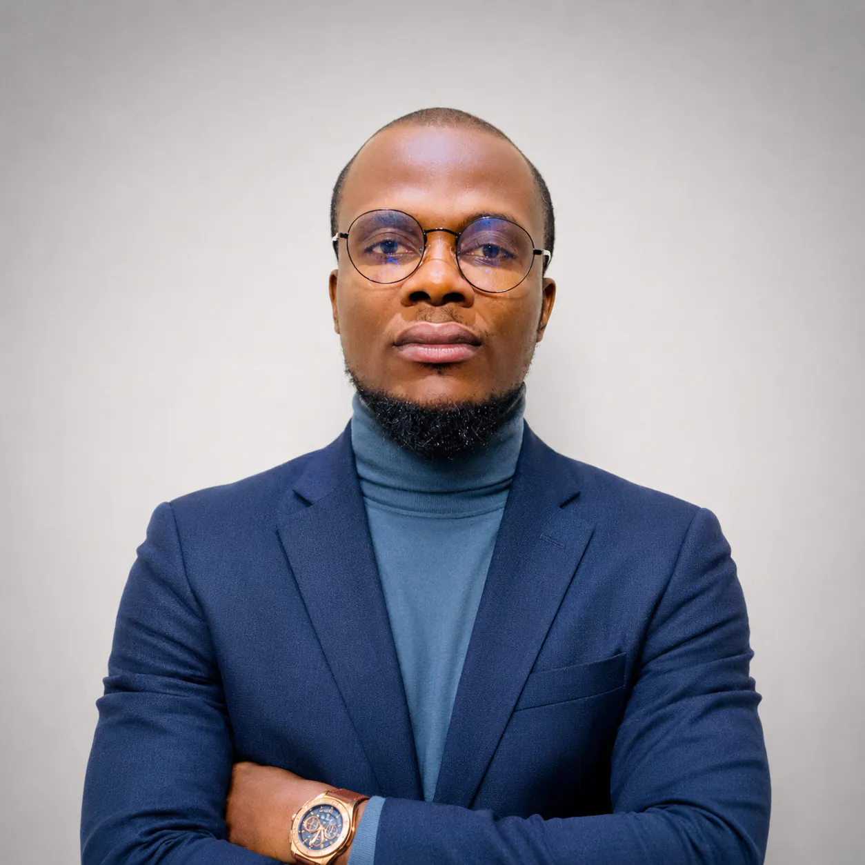 Chukwuma Ukaha, in round wire glasses, a navy blazer and a blue turtleneck, arms folded, looking straight at the camera against a plain grey backdrop.