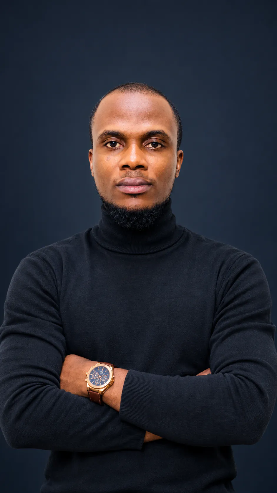 Chukwuma Ukaha, in a black turtleneck with his arms folded, against a dark studio backdrop.